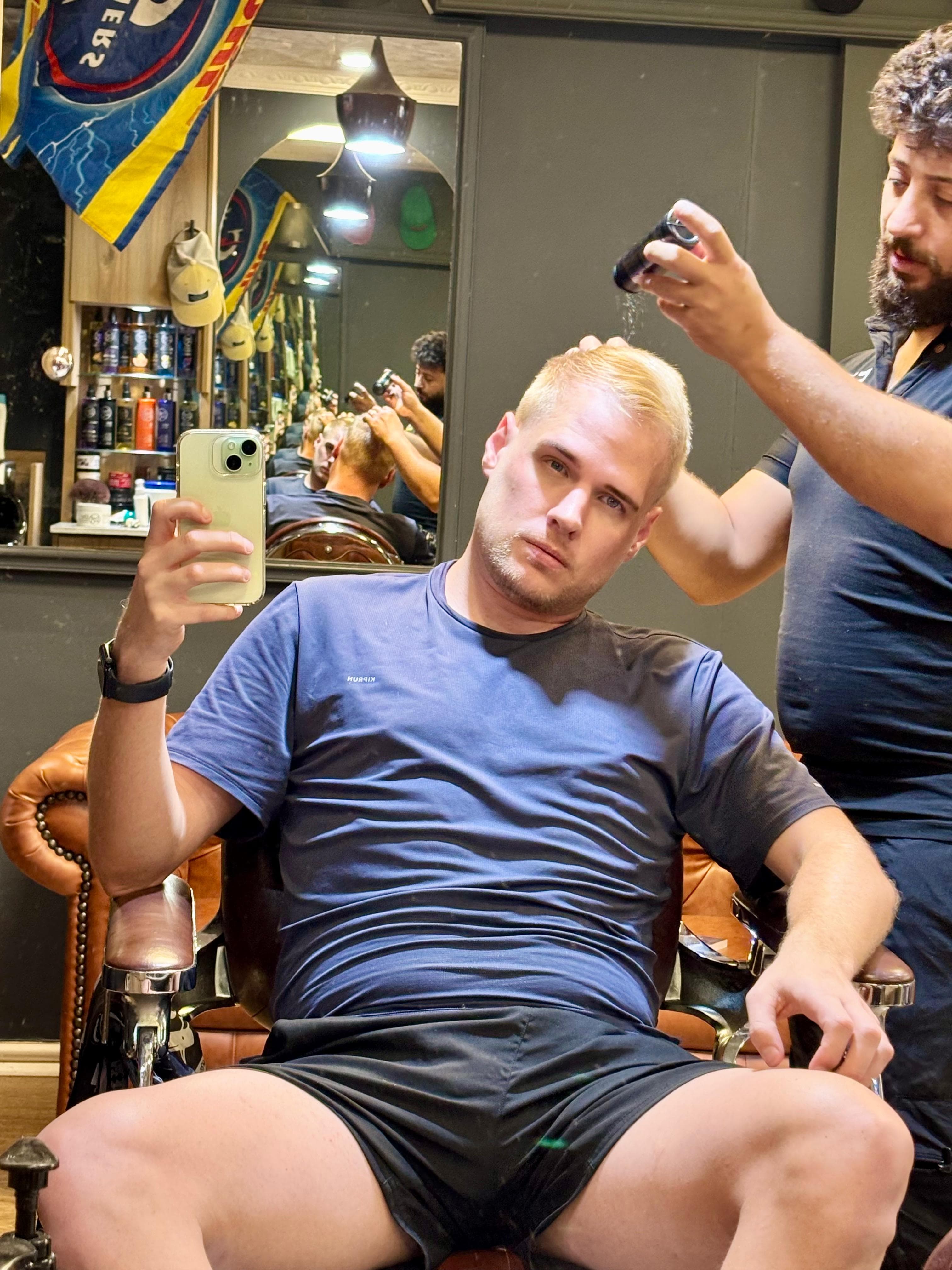 Blonde man taking a selfie while a barber applies styling powder to his hair.