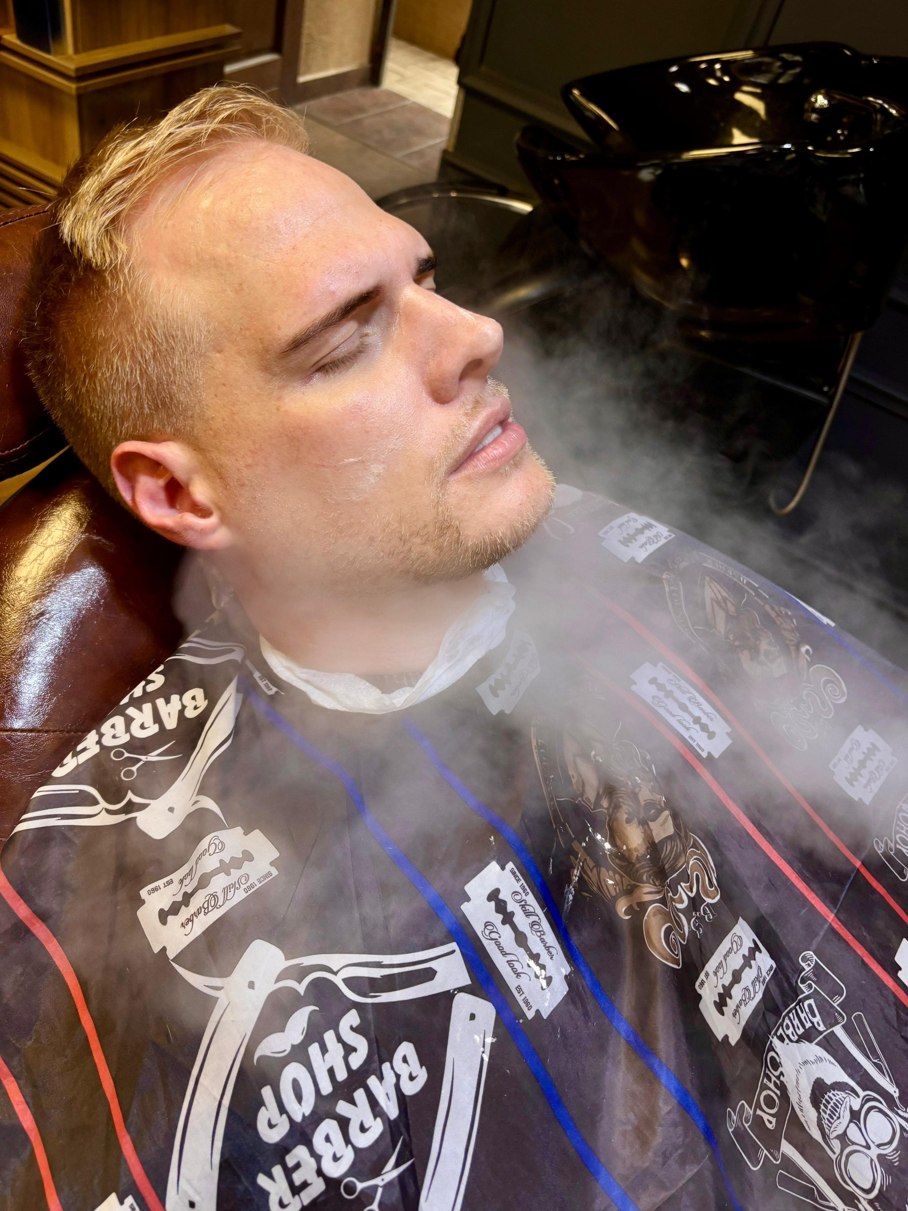 Man with closed eyes reclining in a barber chair, receiving a facial steam treatment.