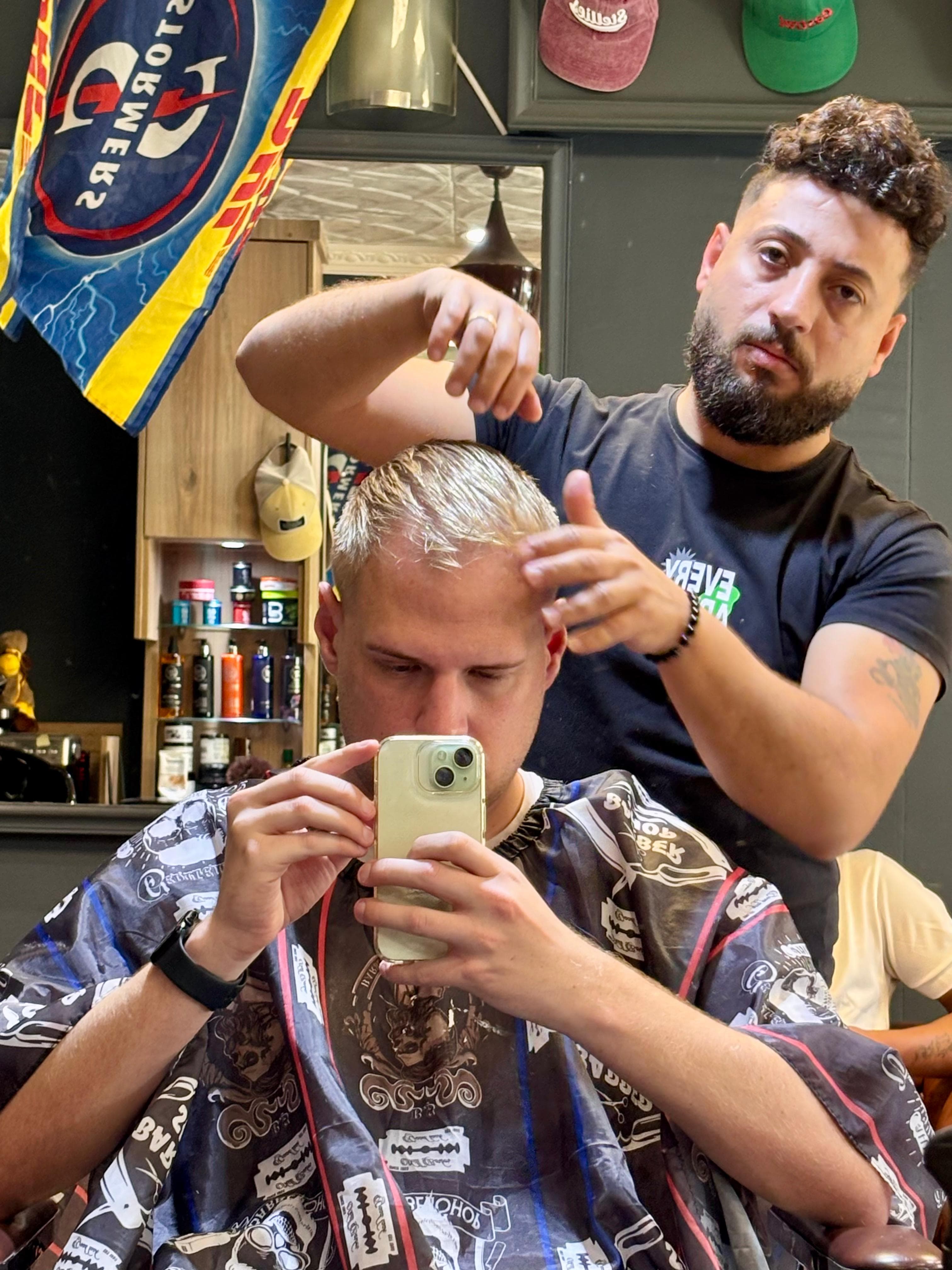 Man takes a mirror selfie while a barber styles his blonde hair in a barbershop.