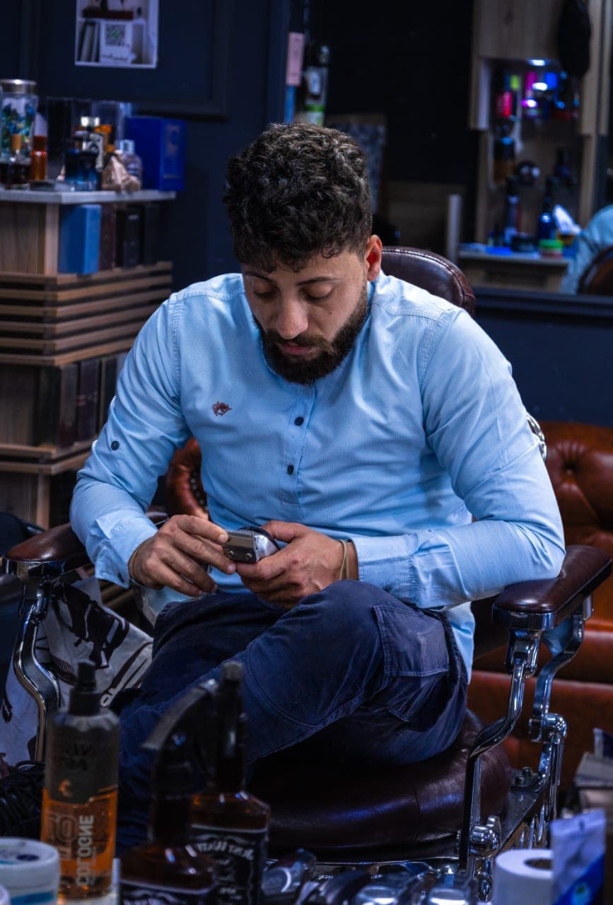 Bearded man in a blue shirt sits in a barber chair, inspecting electric hair clippers.