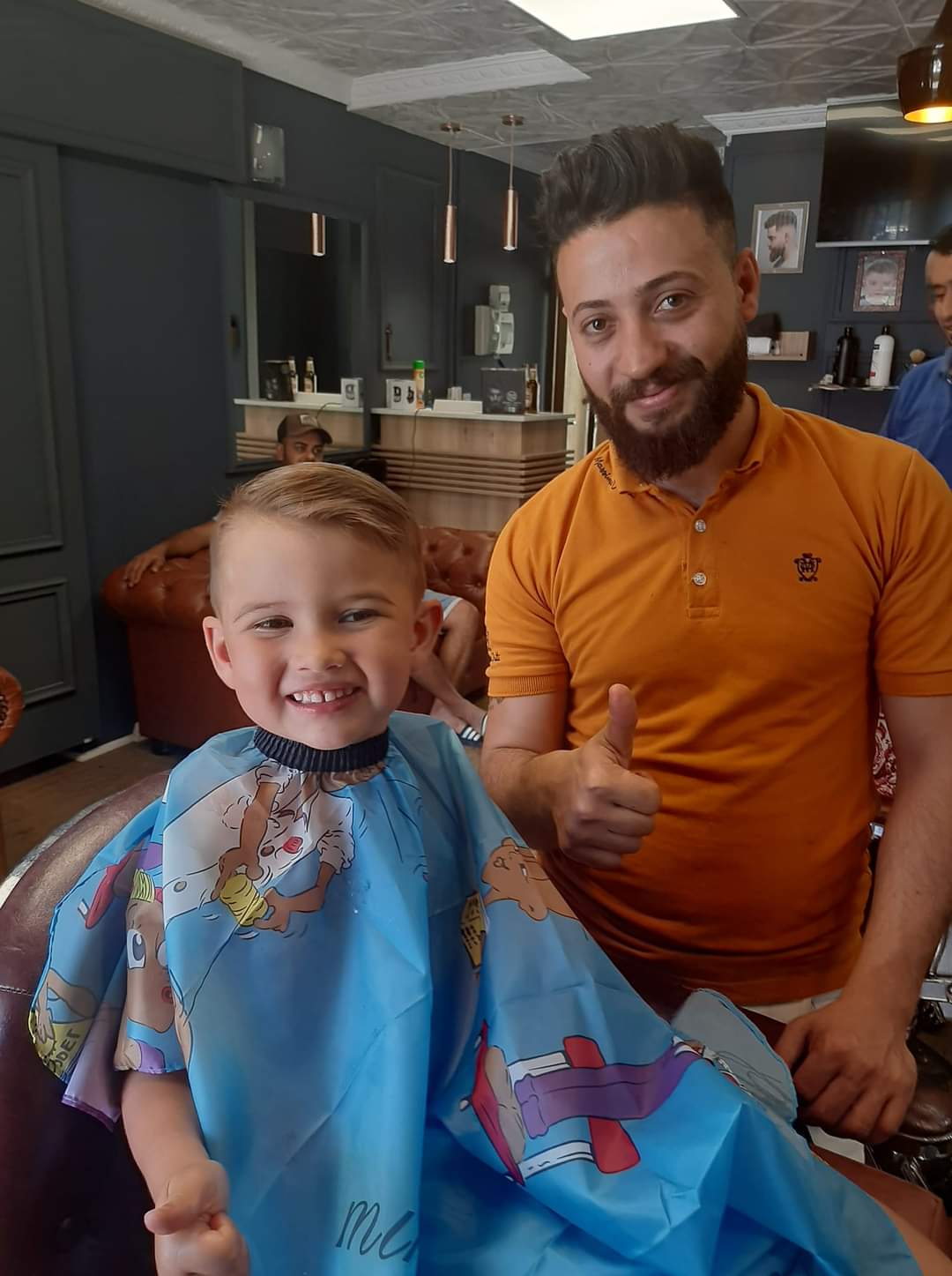 Smiling boy with a fresh haircut sits in a barber chair beside a bearded barber.