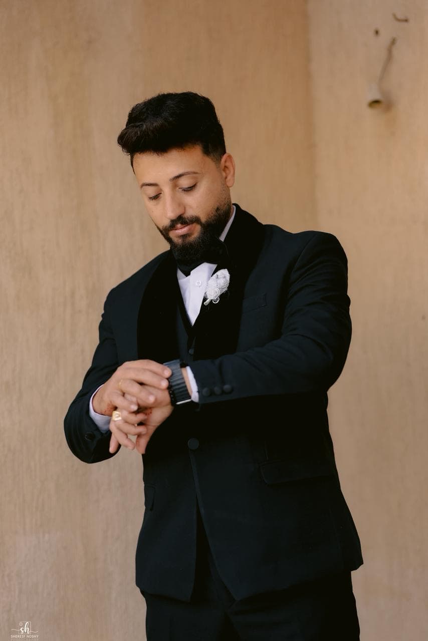 Bearded man in a black tuxedo checks his watch against a textured beige wall.