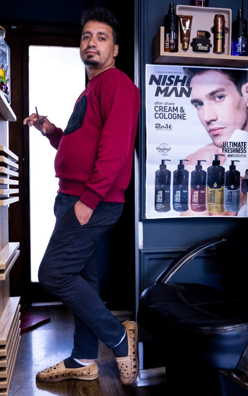 Barber in a red sweater holds scissors, standing in a barbershop next to grooming posters.