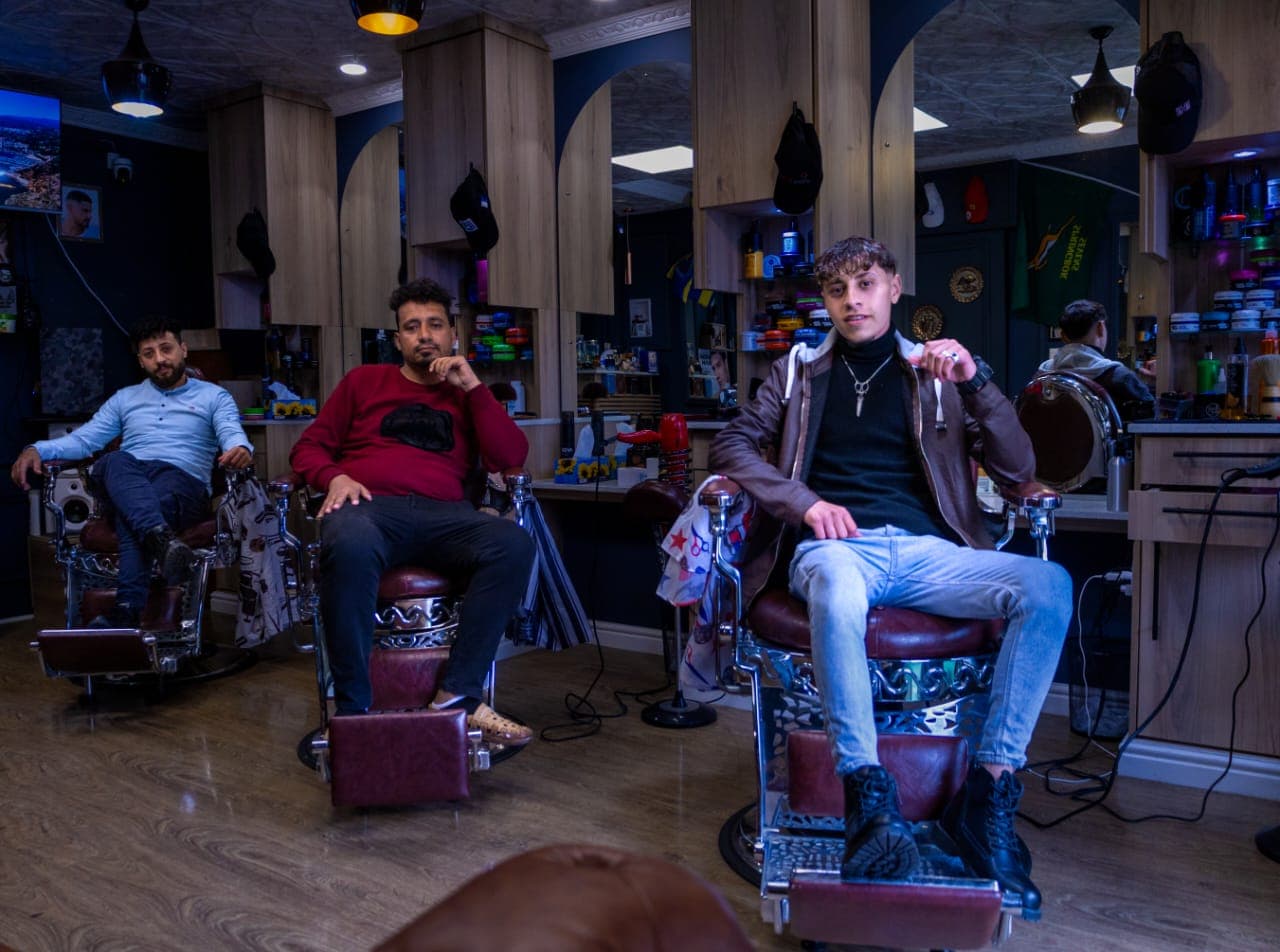 Three men sitting in classic barber chairs inside a barbershop with wooden cabinets and mirrors.